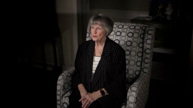 An elderly woman sitting thoughtfully in a patterned armchair during her biography film interview.